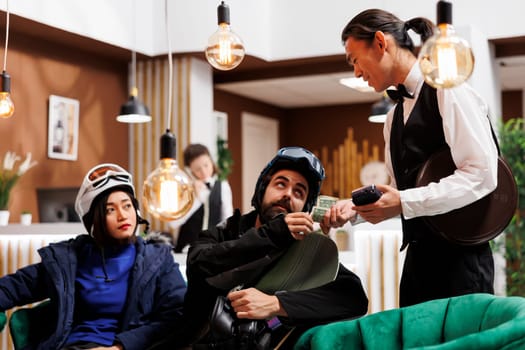 Friendly asian waiter receiving money tip from couple with snow clothing in lounge area of ski mountain resort. Healthy boyfriend and girlfriend thanking employee for good service in winter vacation.