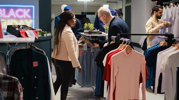 Diverse clients shopping in retail store, looking for black friday discounts. Customers browsing through clothing items on hangers and racks in fashion outlet, exploring seasonal sales.