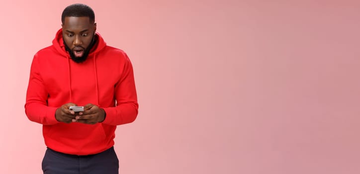 Amused focused impressed african-american bearded guy in red hoodie drop jaw widen eyes look amazed smartphone display winning hard game level answering exciting message, standing pink background.