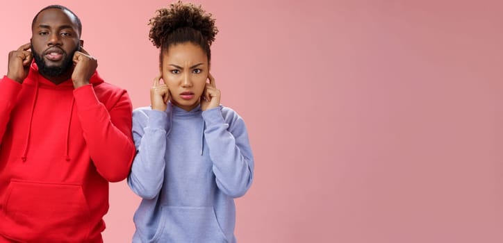 Annoyed tired couple african american woman man relationship exhausted each other close ears plug index fingers frowning look irritated hear terrible disturbing noise, standing pink background.