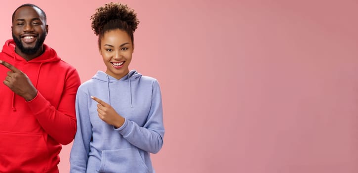 Charming carefree smiling african american two man woman grinning white teeth having fun fool around together pointing upper left corner showing couple project proudly, standing pink background.