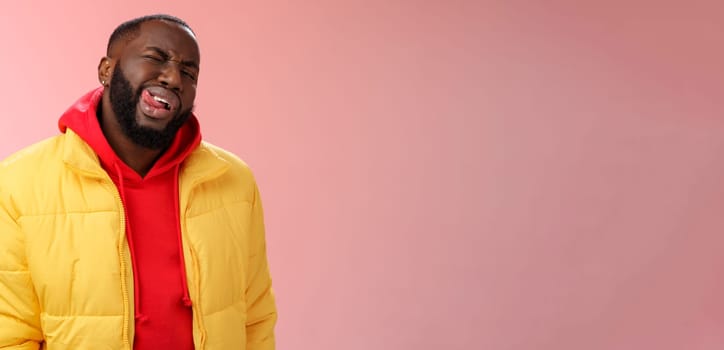 Cheeky stylish funny african bearded man in yellow trendy jacket red hoodie show tongue sassy flirty gaze winking camera trying impress woman pretend macho, standing pink background.