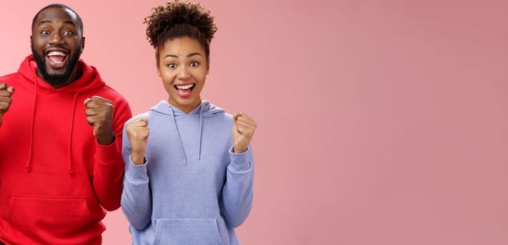 Lucky cheerful two african-american man woman yelling hooray celebrating triumphing huge success clenching fists joyfully accomplish mutual goal standing joyfully pink background victory gesture.