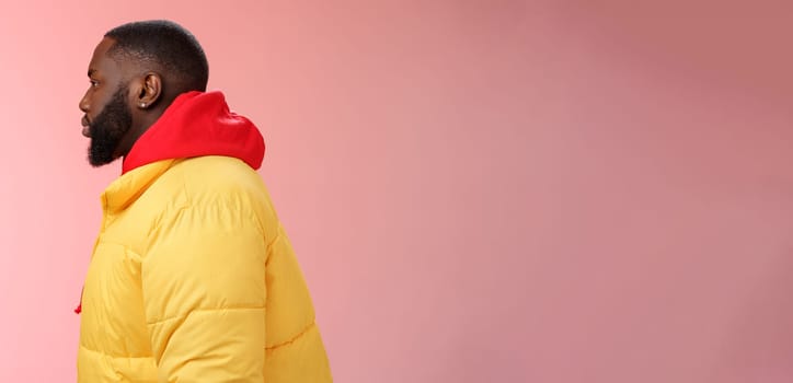 Profile studio shot bearded young 25s african guy in yellow jacket red hoodie look left normal unbothered relaxed expression standing queue order fastfood pink background, waiting take-away.