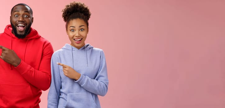 Impressed two best friends african american man woman 25s drop jaw amused visit awesome interesting park pointing left index fingers widen eyes thrilled having fun together, standing pink background.