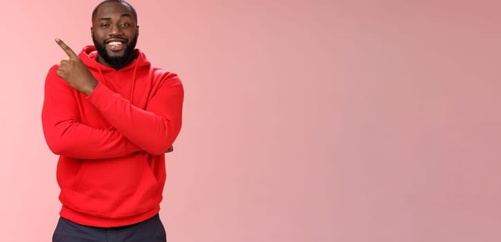 Cheeky outgoing smiling attractive african-american male model in red hoodie grinning energized pointing upper left corner indicating awesome product recommend promo, standing pink background.