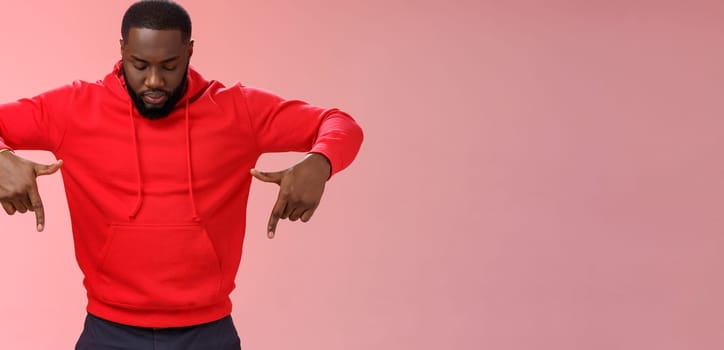 Focused interested serious-looking young african american bearded strong guy wearing red hoodie bend head look pointing down curiously, intrigued what happening lower floor, standing pink background.