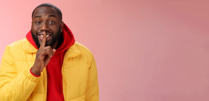 Lifestyle. Charming happy young african-american bearded guy in yellow jacket red hoodie prepare lovely birthday surprise smiling show shush shh gesture index finger on mouth promise keep secret.