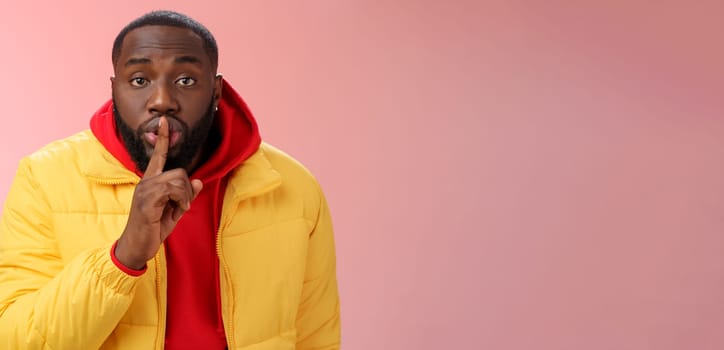 Waist-up serious-looking african-american bearded man in yellow jacket red hoodie bend camera asking keep quiet say shhh show shush gesture index finger on lips, confidential information taboo.