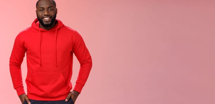 Handsome young happy black bearded guy white perfect smile hold hands pockets casually standing pink background talking have interesting nice conversation, wearing red hoodie. Copy space