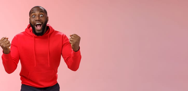 Very happy lucky black bearded guy winning lottery made winning bet clench fists triumphing yelling yes widen eyes surprised joyfully celebrating good news standing impressed, pink background.