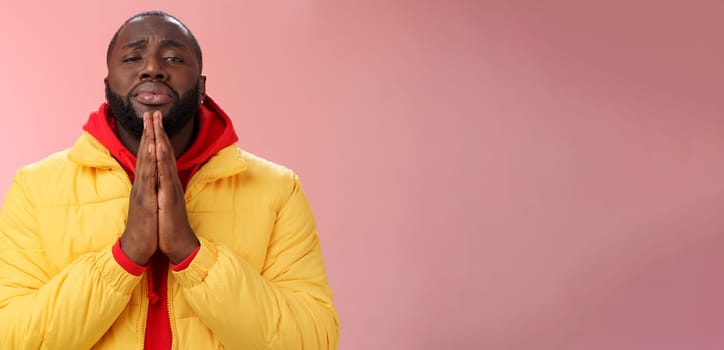 Miserable cute african-american bearded man in yellow coat asking help begging press palms supplicating apologizing please help, standing pink background sad need advice lending money.