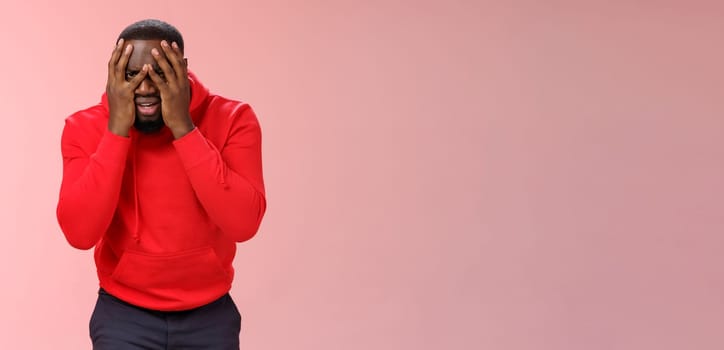 Shocked upset young african american bearded guy in red hoodie facepalm hide face hands peeking through fingers sad devastated lose hope, standing sad pink background grieving feel sorrow.