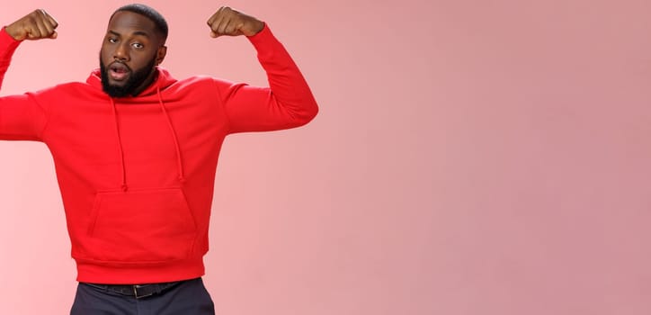 Strong cheeky serious-looking african american sportsman in red hoodie bragging showing-off raise hands excercising show big muscles biceps, standing confident pink background. Copy space