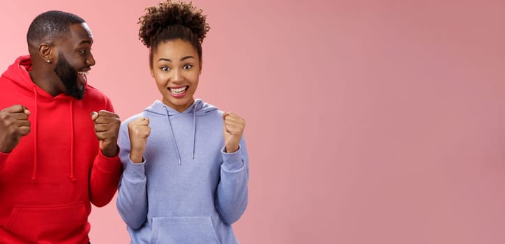 Happy enthusiastic thrilled young african american couple clenching fists joyfully surprised winning awesome honeymoon trip tickets standing pink background triumphing smiling broadly celebrating.