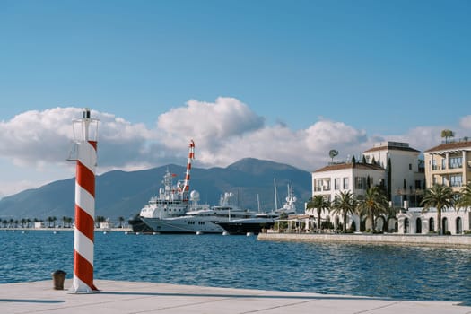 Small striped lighthouse stands on the pier of a luxury marina with moored yachts. High quality photo