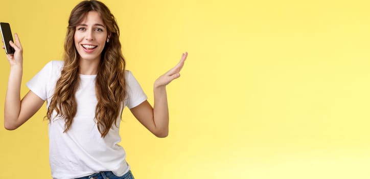 Carefree good-looking lively sociable curly-haired girl having fun wearing wireless earbuds dancing joyfully listening music moving rhythm song hold smartphone raising hands up smiling camera amused. Lifestyle.