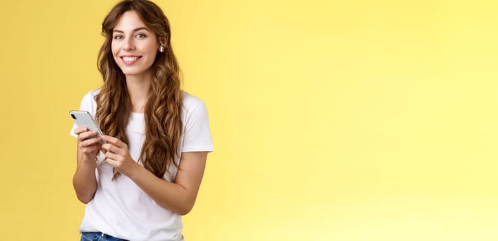 Feminine lovely curly-haired woman white t-shirt hold smartphone picked awesome new song listen music wearing wireless earphones smiling delighted camera enjoy earbuds beats yellow background. Lifestyle.