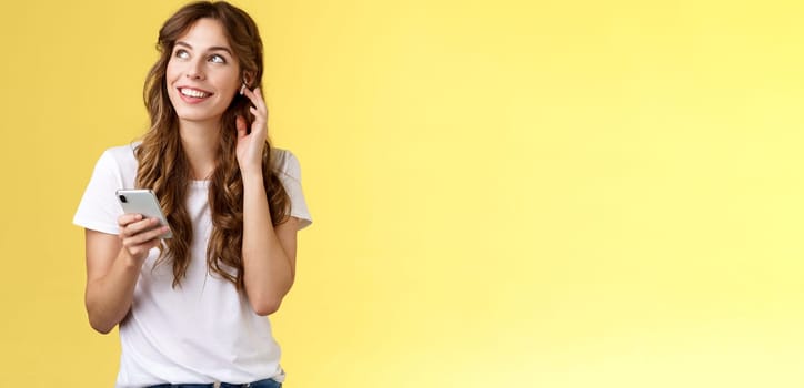 Dreamy happy cheerful curly-haired girl look around contemplate beautiful summer weather listening music touch wireless earbud calling friend talking via earphones hold smartphone yellow background. Lifestyle.