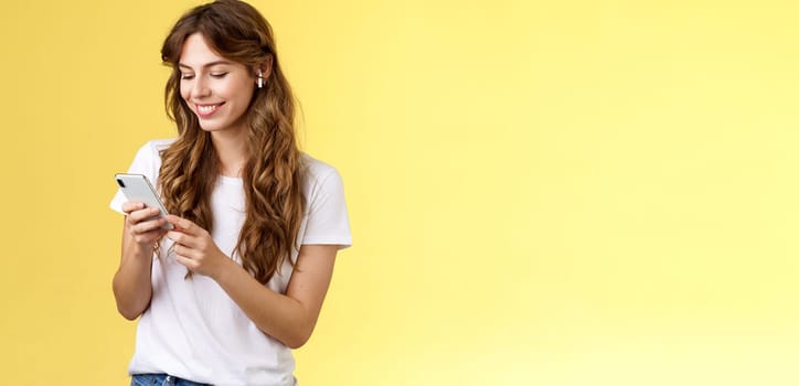 Happy caring tender feminine girlfriend curly long hair hold smartphone picking song listen way home smiling broadly look mobile phone scroll music platform wear wireless earbud yellow background. Lifestyle.