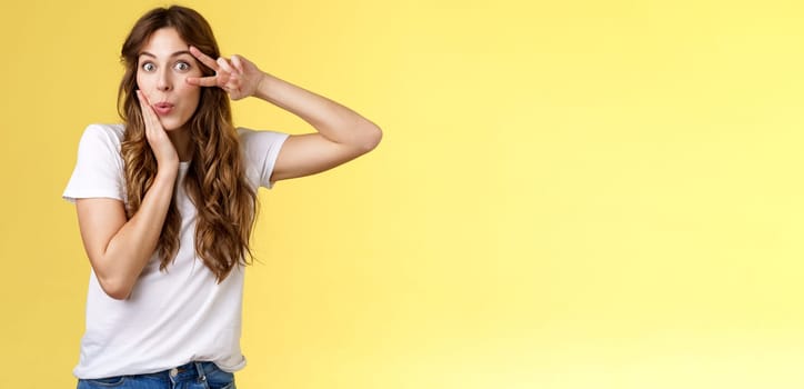 Silly coquesttish good-looking caucasian girl curly hairstyle folding lips amused interested touch cheek feminine sensual pose show victory peace sign upbeat stand intrigued yellow background. Lifestyle.