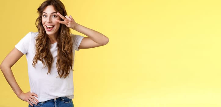Playful surprised charismatic young caucasian girlfriend having fun look amused enthusiastic show peace victory sign laughing smiling joyfully enjoy summer holidays touch hip stand yellow background. Lifestyle.