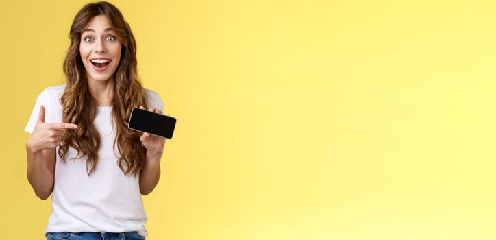 Cheerful surprised cute lucky girl beat best score awesome game showing smartphone display pointing index finger horizontal screen mobile phone introduce cool app smiling broadly yellow background. Lifestyle.