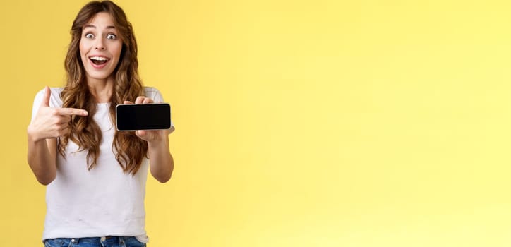 Impressed upbeat happy lucky girl curly long hairstyle open mouth admiration joy like awesome new app show smartphone screen horizontal phone display stand yellow background amazed. Lifestyle.