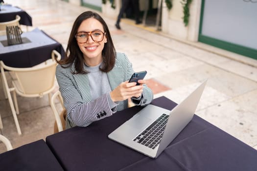 Brunette woman in casual clothes working with laptop using mobile phone at cafe. Freelancer using smartphone and laptop sitting outdoor cafe looking at camera. High angle view, copy space