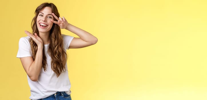 Lively enthusiastic attractive carefree woman enjoy relaxed weekends summer holiday have fun smiling broadly tilt head show peace victory sign joyfully introduce herself posing yellow background. Lifestyle.