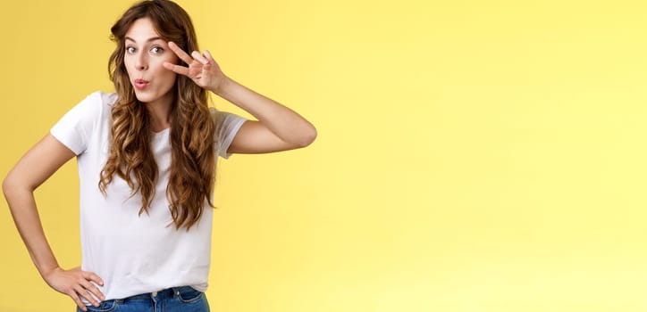 Cute glamour young woman curly haircut tilt head silly show peace victory sign fold lips lovely muah kiss gesture send flirty glances camera hold hip mimicking sensually gazing you yellow background. Lifestyle.