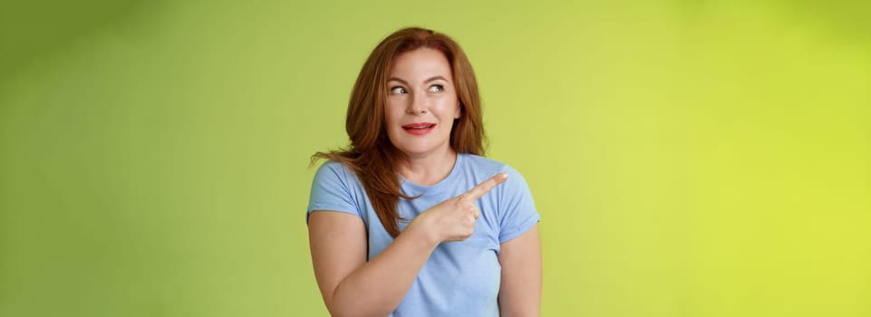 Excited intrigued middle-aged mature redhead woman. pointing gazing left side copy space curiously smiling thrilled like interesting promo willing check-out great advertisement green background.