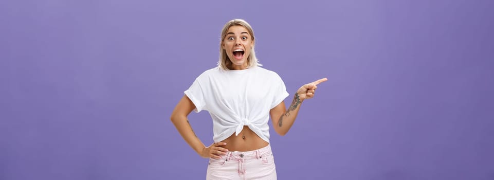 Thrilled and excited positive attractive woman with fair hair tanned skin and tattooed arms smiling joyfully with opened mouth staring impressed and delighted at camera, pointing left over purple wall.