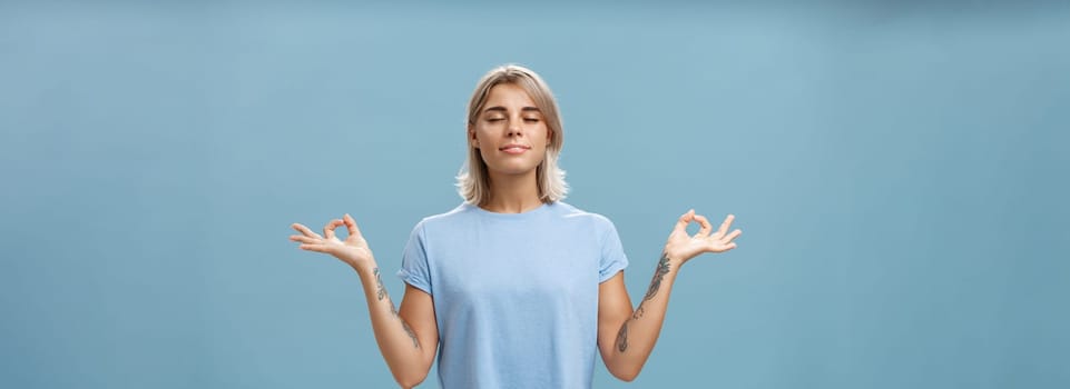 Listening to inner self during meditation. Calm happy and relaxed attractive tanned blonde female with tattoos on arms standing in lotus pose over blue wall with zen gesture closed eyes doing yoga.