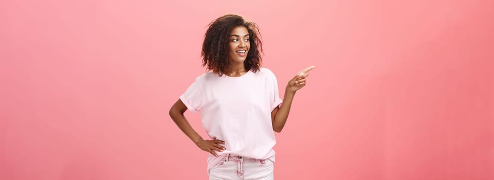 Girl checking out nice body of guy passing by. Charming confident dark-skinned female with curly hairstyle holding hand on hip looking and pointing left discussing cool copy space over pink wall.