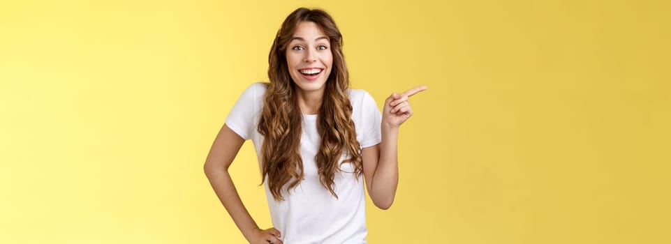 Lively surprised happy enthusiastic good-looking woman long curly hairstyle white t-shirt laughing impressed astonished pointing left index finger discuss interesting exhibition yellow background.