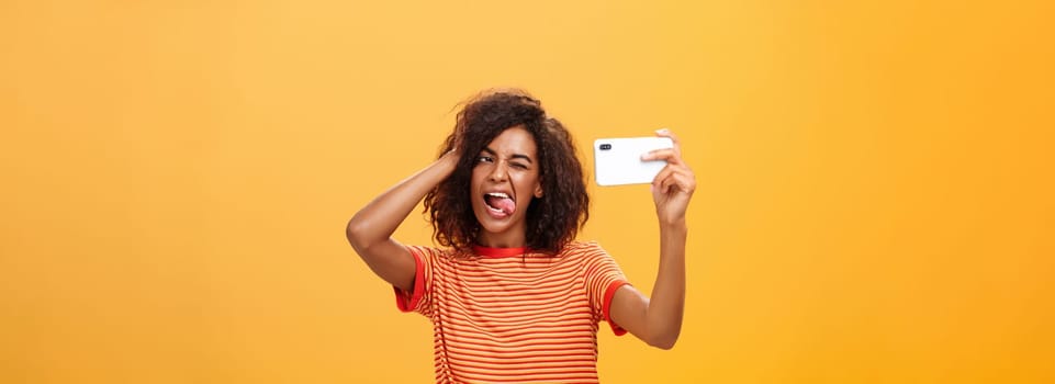 Waist-up shot of stylish confident african american woman with afro hairstyle in trendy t-shirt taking selfie on awesome new cellphone sticking out tongue playing with hair and winking at smartphone. Technology and people concept