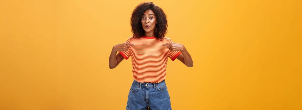 Woman being delighted telling friend she got new job pointing at herself with pleased satisfied look bragging standing happy over orange background in stylish denim shorts and striped t-shirt. Lifestyle.