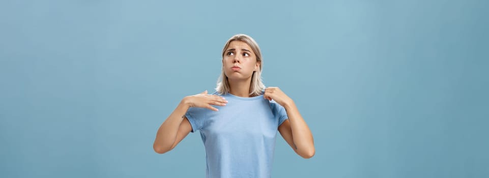Girl feeling discomfort from heat standing over blue background in fug breathing out and frowning looking up at sun suffering from hot weather waving with t-shirt to cool. Body language concept