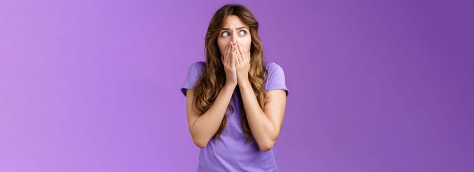 Shocked timid insecure concerned curly-haired woman look sideways stunned scared gasping cover mouth hold palms pressed face stare left frightened troubled panic purple background.