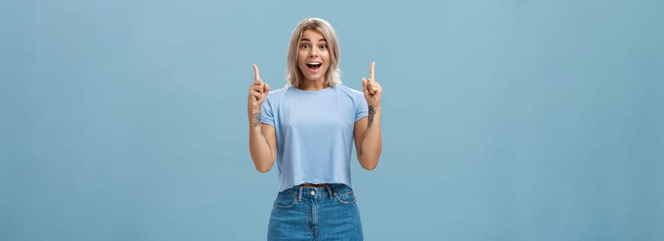 Waist-up shot of impressed enthusiastic creative blonde woman in trendy summer outfit smiling gasping being charmed and thrilled with awesome copy space pointing up over blue background. Copy space