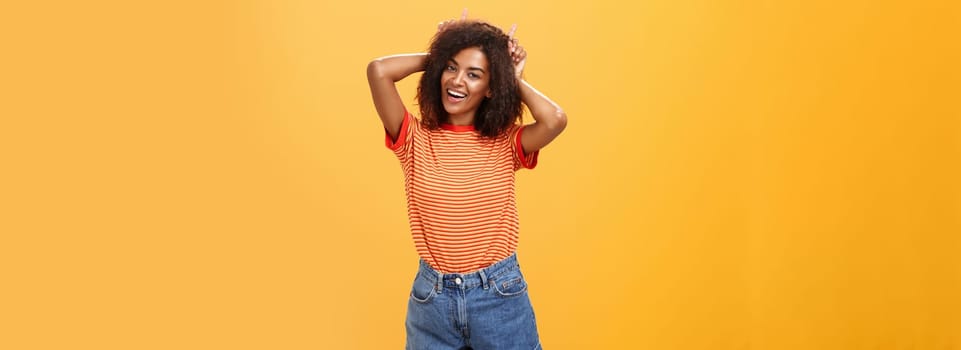 Rebellious playful and joyful african american girl with curly hairstyle making devil horns on head with index fingers smiling broadly showing daredevil character and charisma over orange wall. Lifestyle.