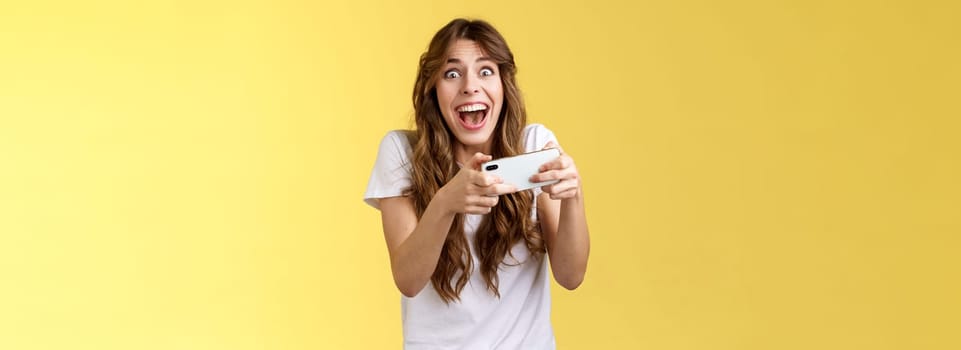 Extremely happy thrilled playful girl gamer playing awesome great new smartphone game hold mobile phone horizontal cheering look camera astonished impressed beating record yellow background.