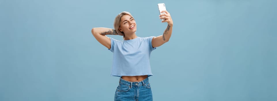 New post in my blog. Stylish feminine and sociable good-looking young female posing for selfie with pulled arm and smartphone sticking out tongue while making faces at device screen over blue wall.