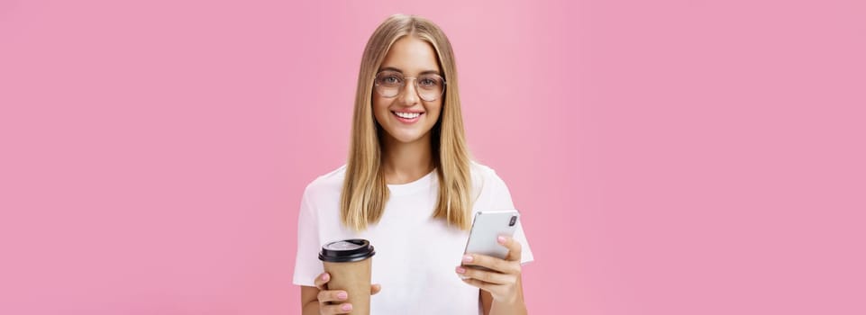 Pleasant friendly-looking girl smiling at camera holding paper cup of coffee and smartphone. Portrait of joyful nice woman drinking morning drink, posing opinion about cafe in internet over pink wall. Technology and lifestyle concept