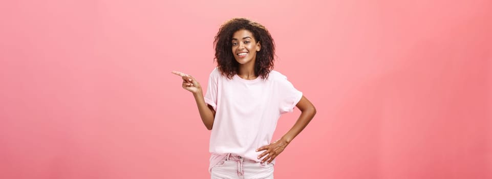 Hey look. Portrait of cute chill and friendly stylish african american woman with afro hairstyle holding hand on waist pointing right and smiling joyfully over pink background. Copy space