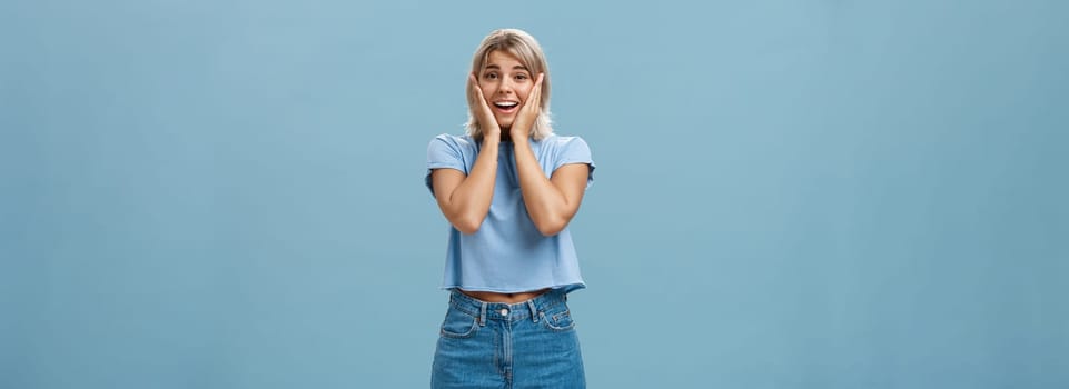 Oh my so cute. Portrait of touched tender good-looking athletic woman with fair hair holding palms on cheeks smiling being impressed and pleased standing in denim shorts over blue background.