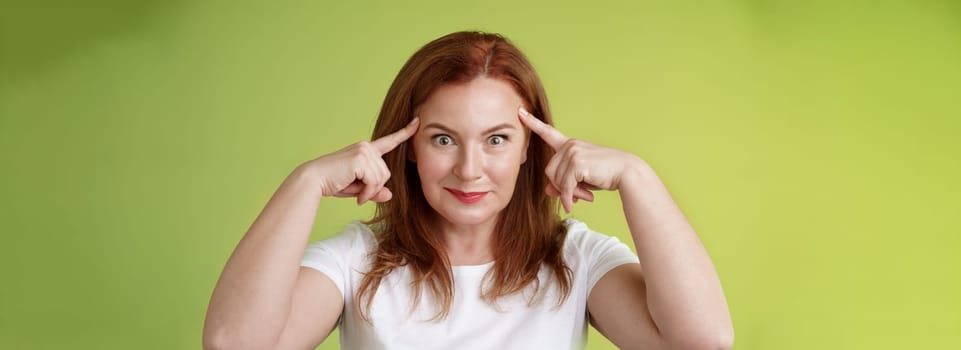 Woman playfully staring funny control your mind. Silly redhead middle-aged female touch temples popping eyes smiling delighted read thoughts trying guess intrigued what thinking green background.