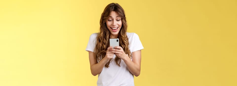 Excited cheerful happy curly-haired silly girl receive surprising good news hold smartphone smiling delighted look mobile phone screen answering b-day invitation stand yellow background upbeat. Lifestyle.