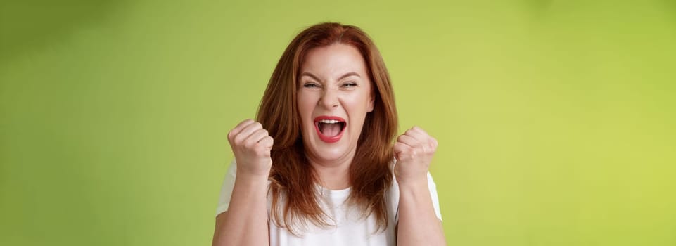 Yeah we did it. Joyful lucky redhead middle-aged female winner pump fists up celebration success gesture yelling triumph joy smiling broadly celebrate awesome news stand green background happy.